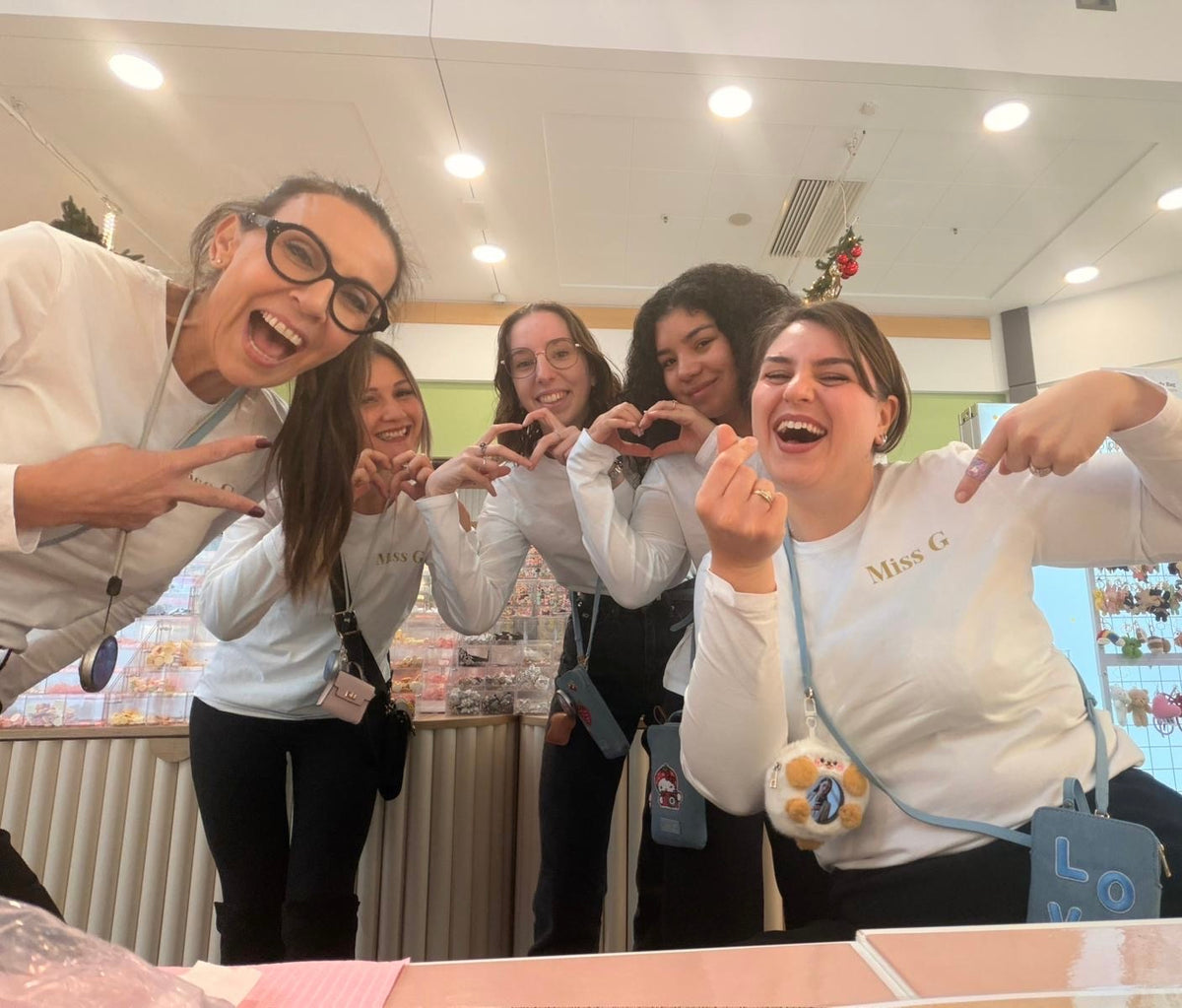 Five women laughing and posing together in a brightly lit boutique or shop interior. Several are wearing white long-sleeve tops, and one has a small crossbody bag with a cartoon face and blue strap.