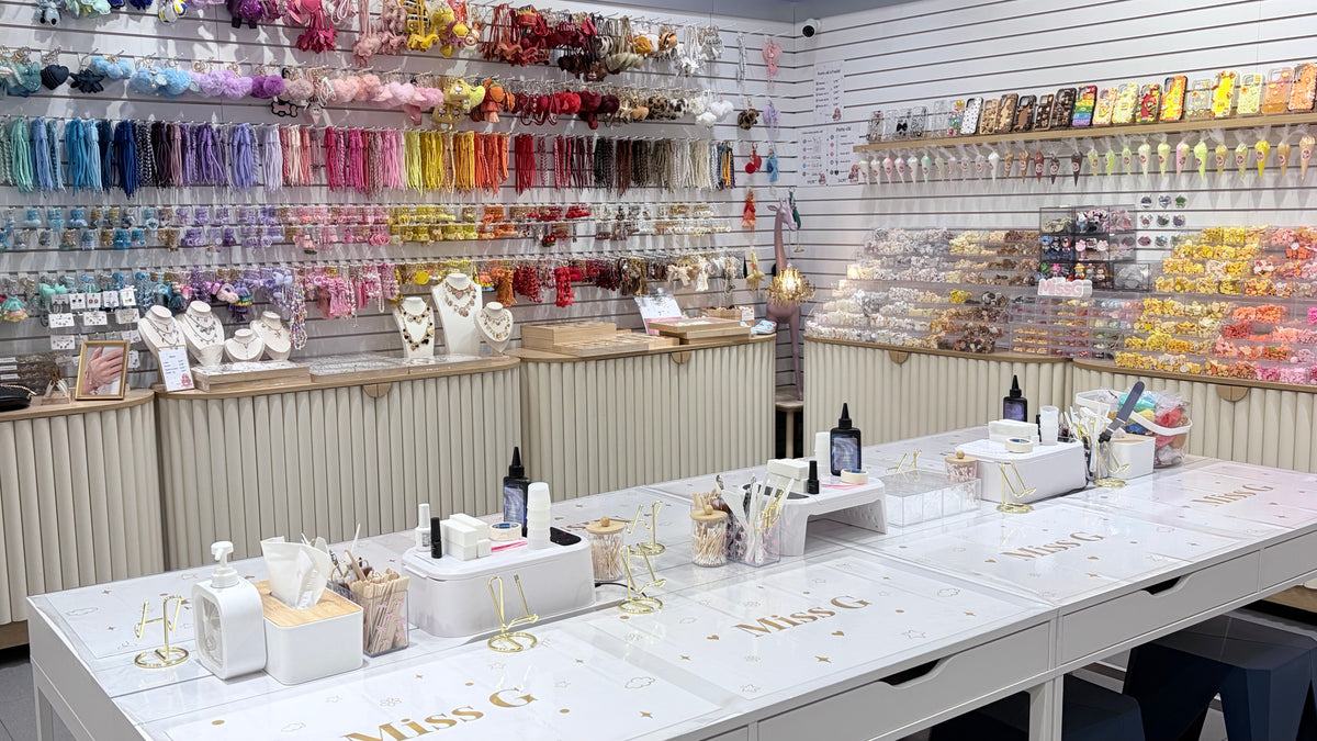 A retail display table inside a cosmetics or beauty store, featuring white drawers with gold lettering, makeup brushes, small bottles, and cosmetic containers. The background is filled with shelves displaying an extensive collection of colorful hair extensions, ribbons, and packaged beauty products.